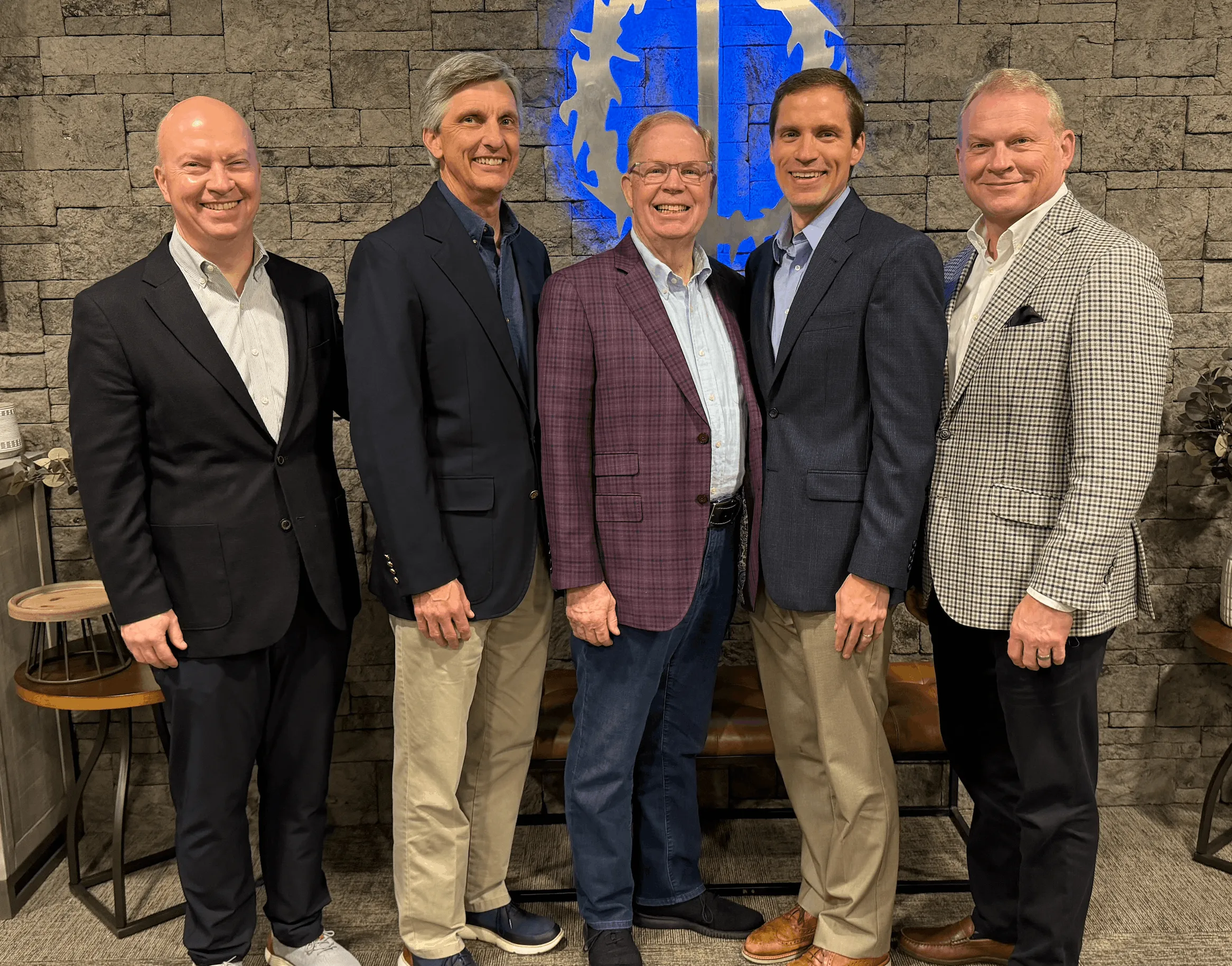Five men dressed in business casual attire standing and smiling in front of a stone wall with a blue circular logo.