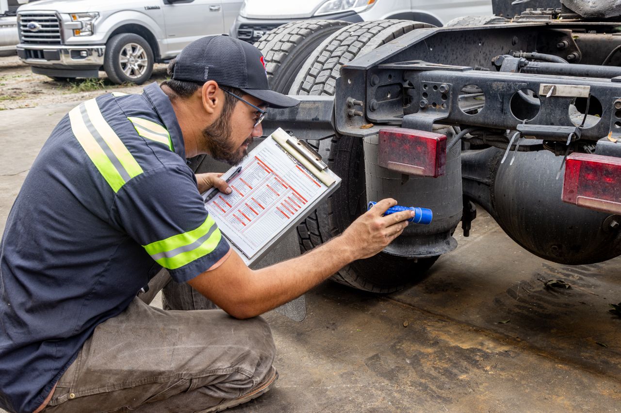 Mechanic kneeling beside a heavy-duty truck, inspecting rear components with flashlight and checklist clipboard during a detailed maintenance and safety inspection.