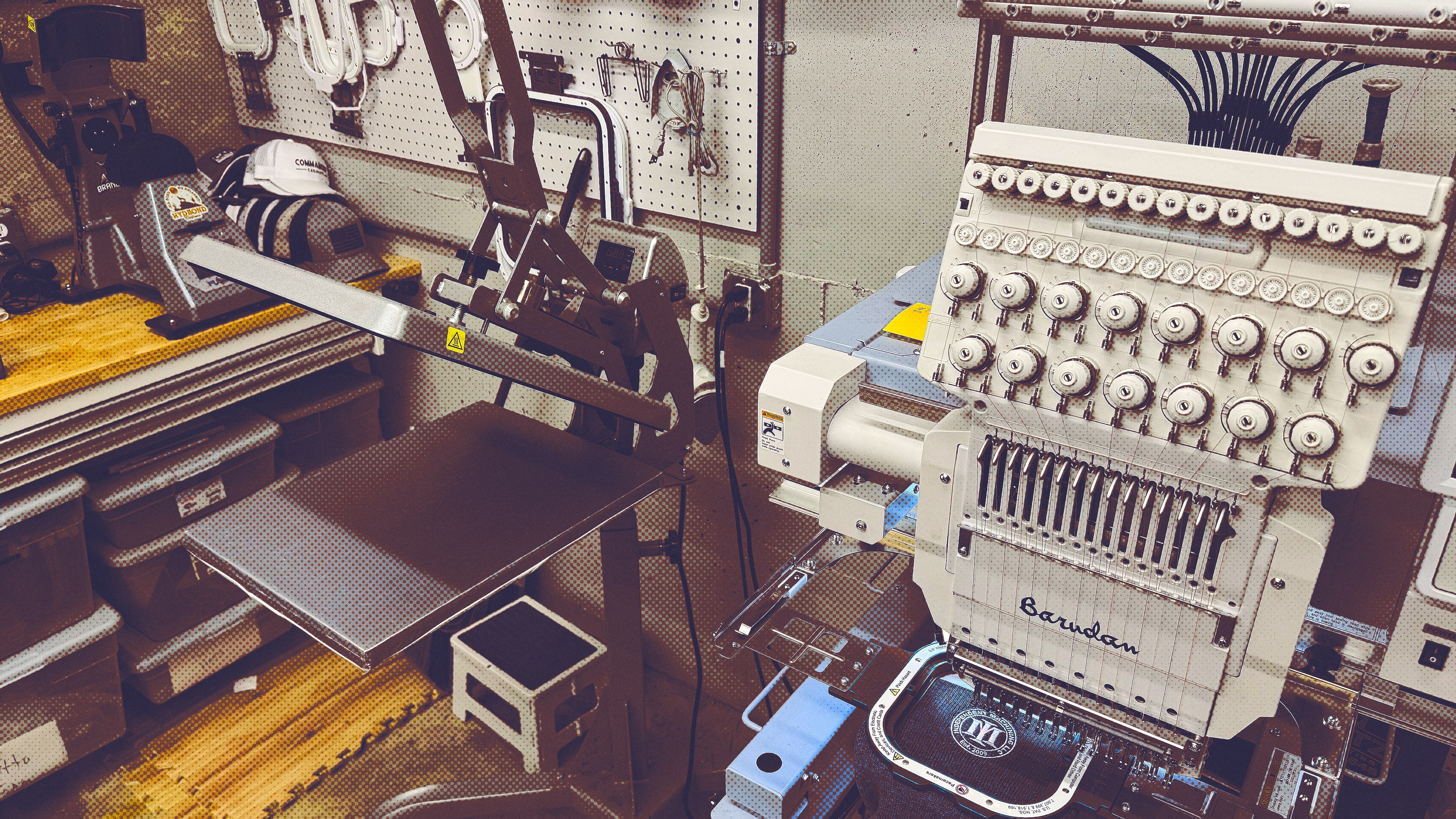 Industrial embroidery machine with multiple thread spools and a heat press machine in a workspace.