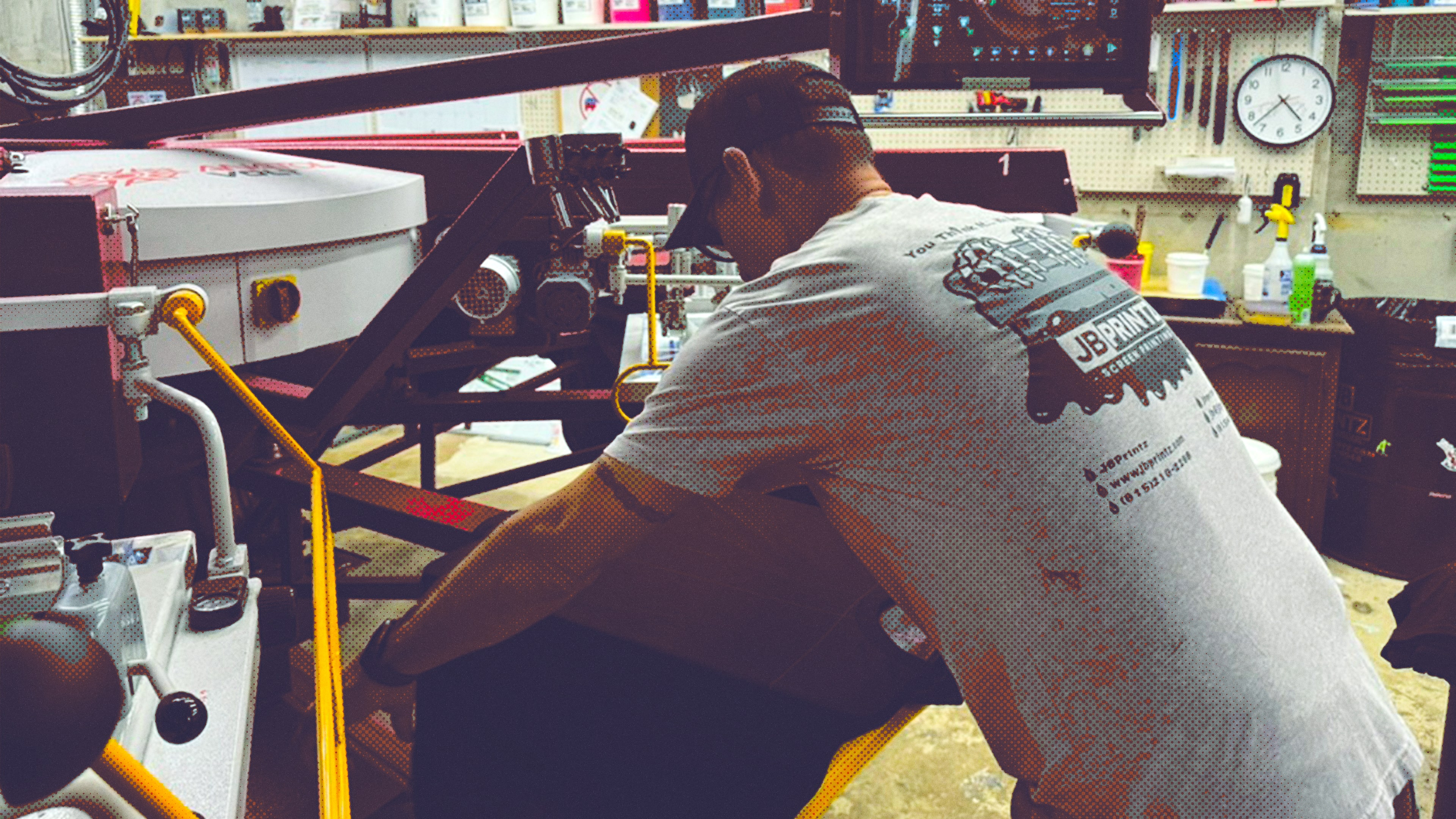 Man operating printing equipment in a workshop wearing a JB Printz t-shirt and a black cap.