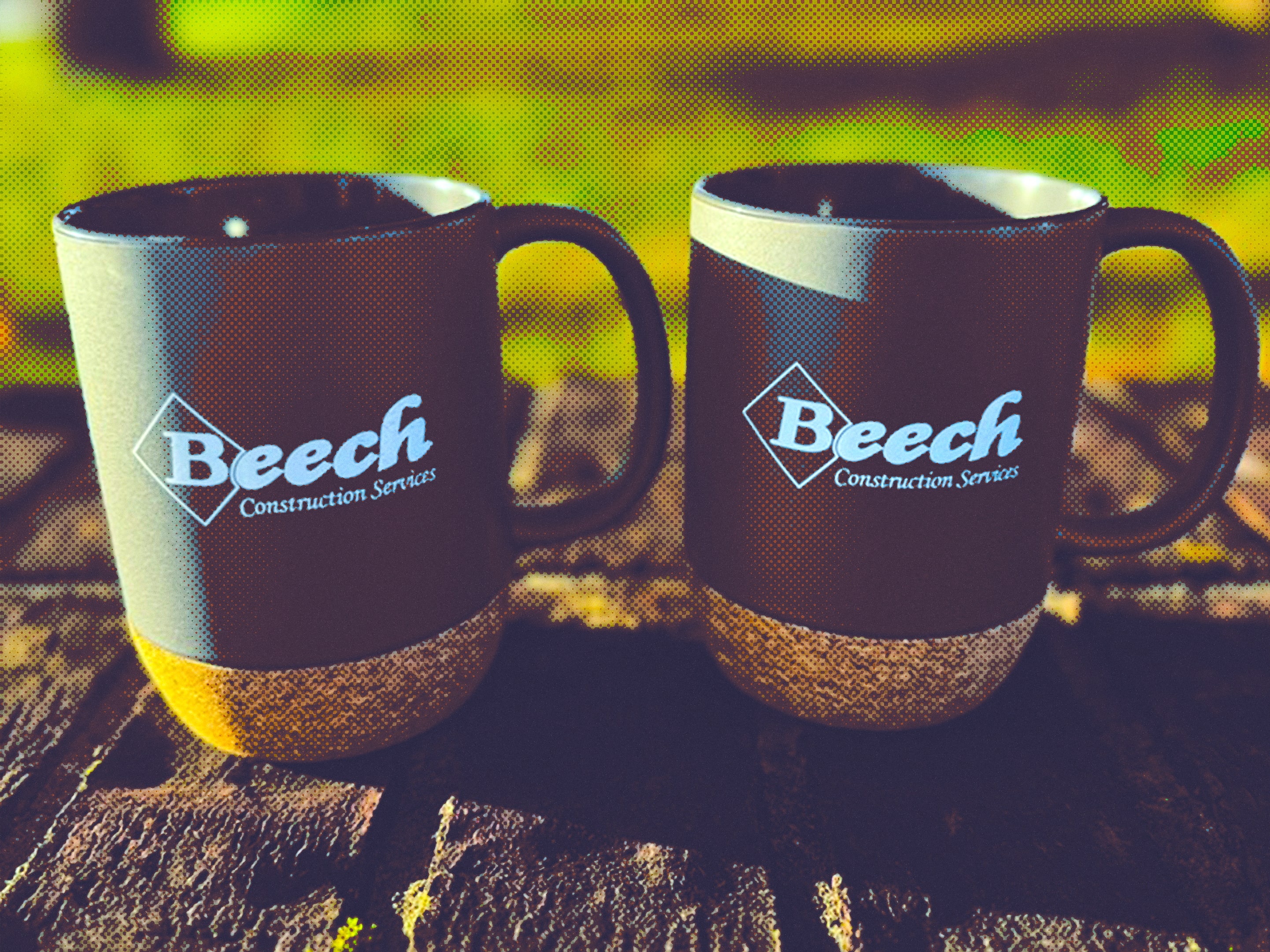 Two dark-colored ceramic mugs with cork bases, each printed with the Beech Construction Services logo, sitting on a wooden surface outdoors.