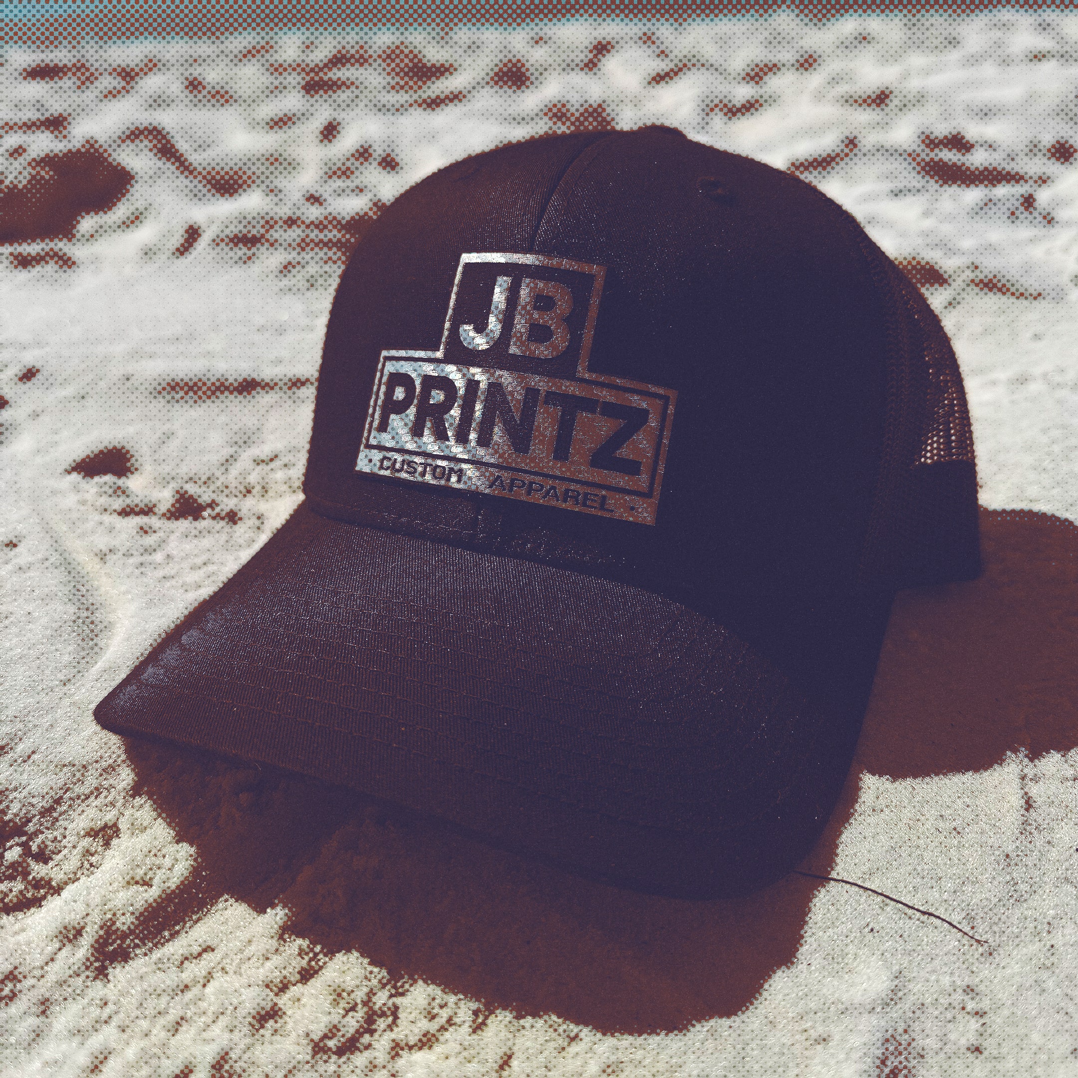 Black trucker hat with silver JB Printz Custom Apparel logo placed on sandy beach with foamy water in the background.