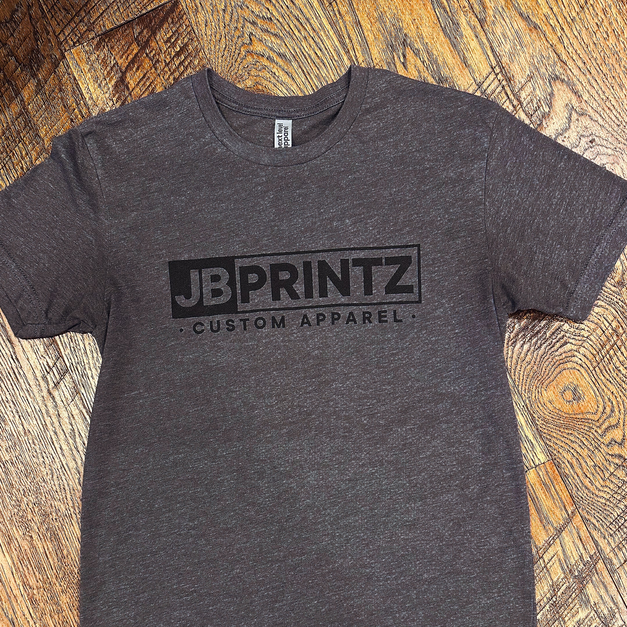 Dark gray t-shirt with JB PRINTZ CUSTOM APPAREL logo printed in black on the front, laid flat on a wooden floor.