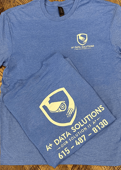 Two blue T-shirts with yellow logos and text for 4+ Data Solutions, featuring a surveillance camera inside a shield with a phone number.