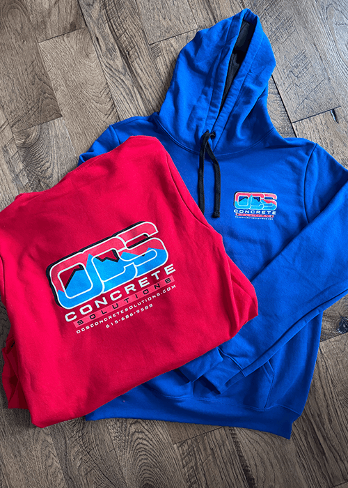 Red and blue hoodies with OCS Concrete Solutions logos placed on a wooden floor.