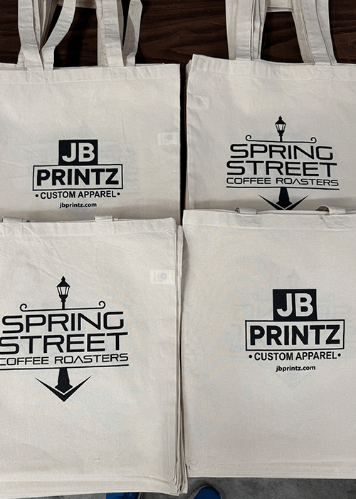 Four beige tote bags laid out on a dark wood surface, two have the logo 'JB PRINTZ CUSTOM APPAREL jbprintz.com' and two have the logo 'SPRING STREET COFFEE ROASTERS' with a street lamp design.