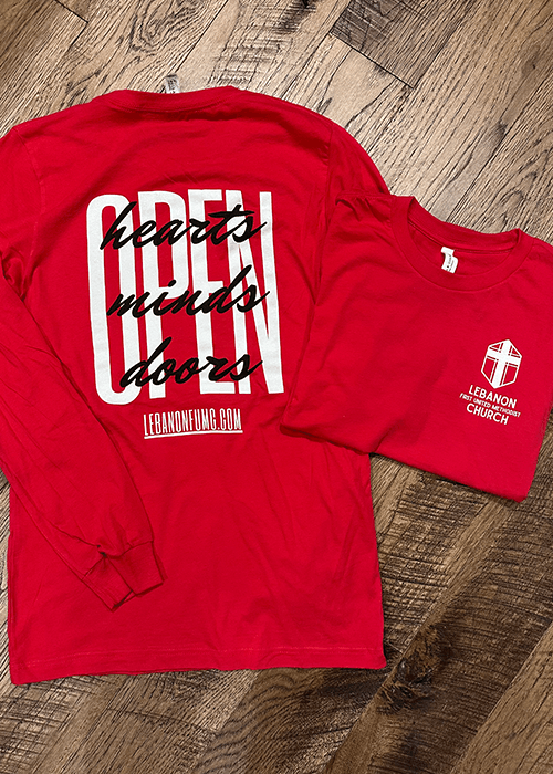 Two red Lebanon First United Methodist Church shirts on a wood floor, one long-sleeve with 'Open hearts minds doors' text and one folded short-sleeve with church logo.
