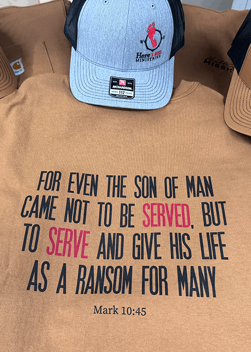 Brown shirt featuring the Bible verse Mark 10:45 in black and red text: 'For even the son of man came not to be served, but to serve and give his life as a ransom for many.' A gray and black cap with a red footprint logo rests on the shirt.
