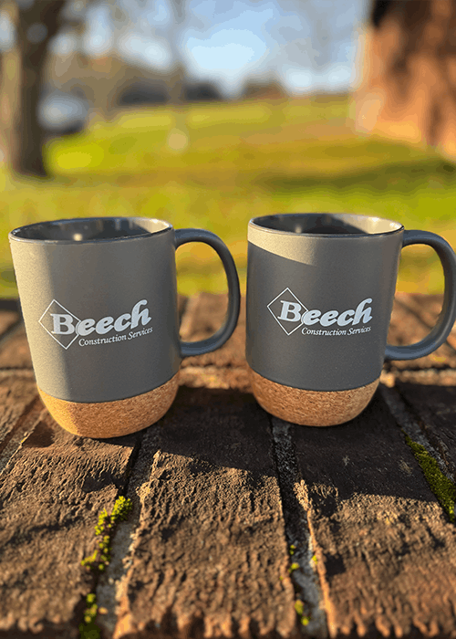Two gray mugs with cork bases on a brick surface, both printed with the Beech Construction Services logo.