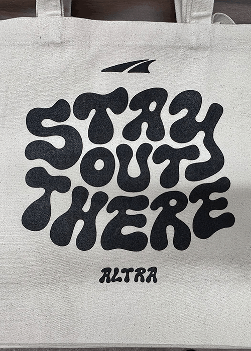 Beige tote bag with black psychedelic-style text reading 'STAY OUT THERE' and smaller text 'ALTRA' below with a simple flag-like logo above.