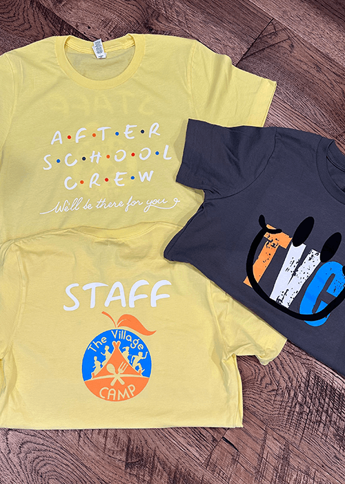 Three T-shirts laid on a wooden floor, two yellow with 'AFTER SCHOOL CREW' and 'STAFF The Village Camp' logos, and one dark grey with a colorful graphic design.