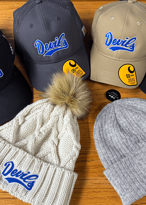 Four hats on a wooden surface: two baseball caps in dark gray and tan with 'Devils' embroidered in blue and white, a cream knit beanie with a fur pom-pom and 'Devils' patch, and a plain light gray ribbed beanie.