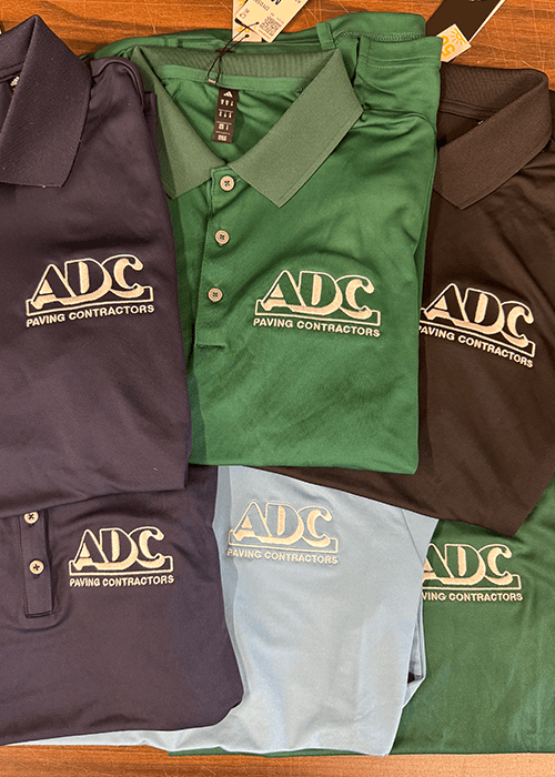 Folded polo shirts in navy, green, black, and light blue colors with ADC Paving Contractors logo embroidered on the chest.