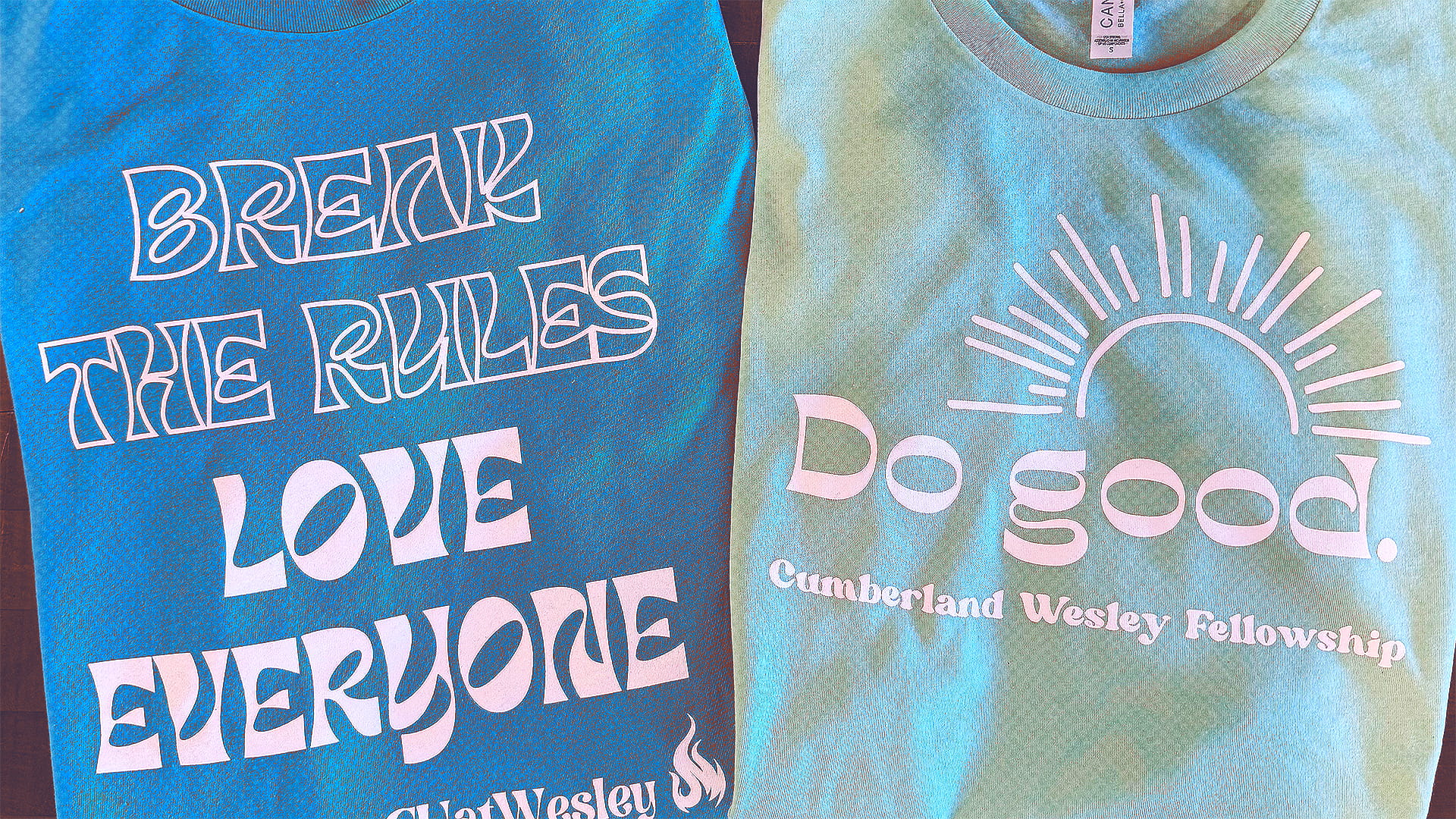 Two folded t-shirts side by side; the left one is blue with white text reading 'BREAK THE RULES LOVE EVERYONE', and the right one is light green with a sun graphic and white text reading 'Do good. Cumberland Wesley Fellowship'.