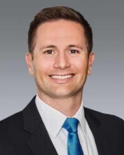 Smiling man in a black suit, white shirt, and blue tie with short brown hair against a gray background.