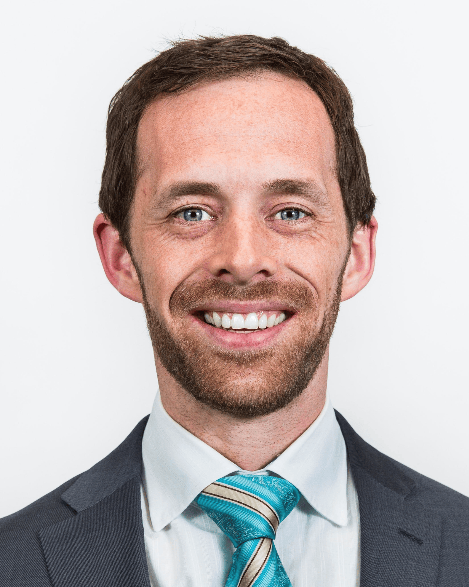 Smiling man with short brown hair, beard, wearing a dark suit, white shirt, and blue striped tie.