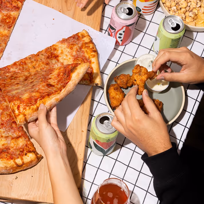 Hands sharing pizza slices, fried chicken, and drinks on a checkered tablecloth.