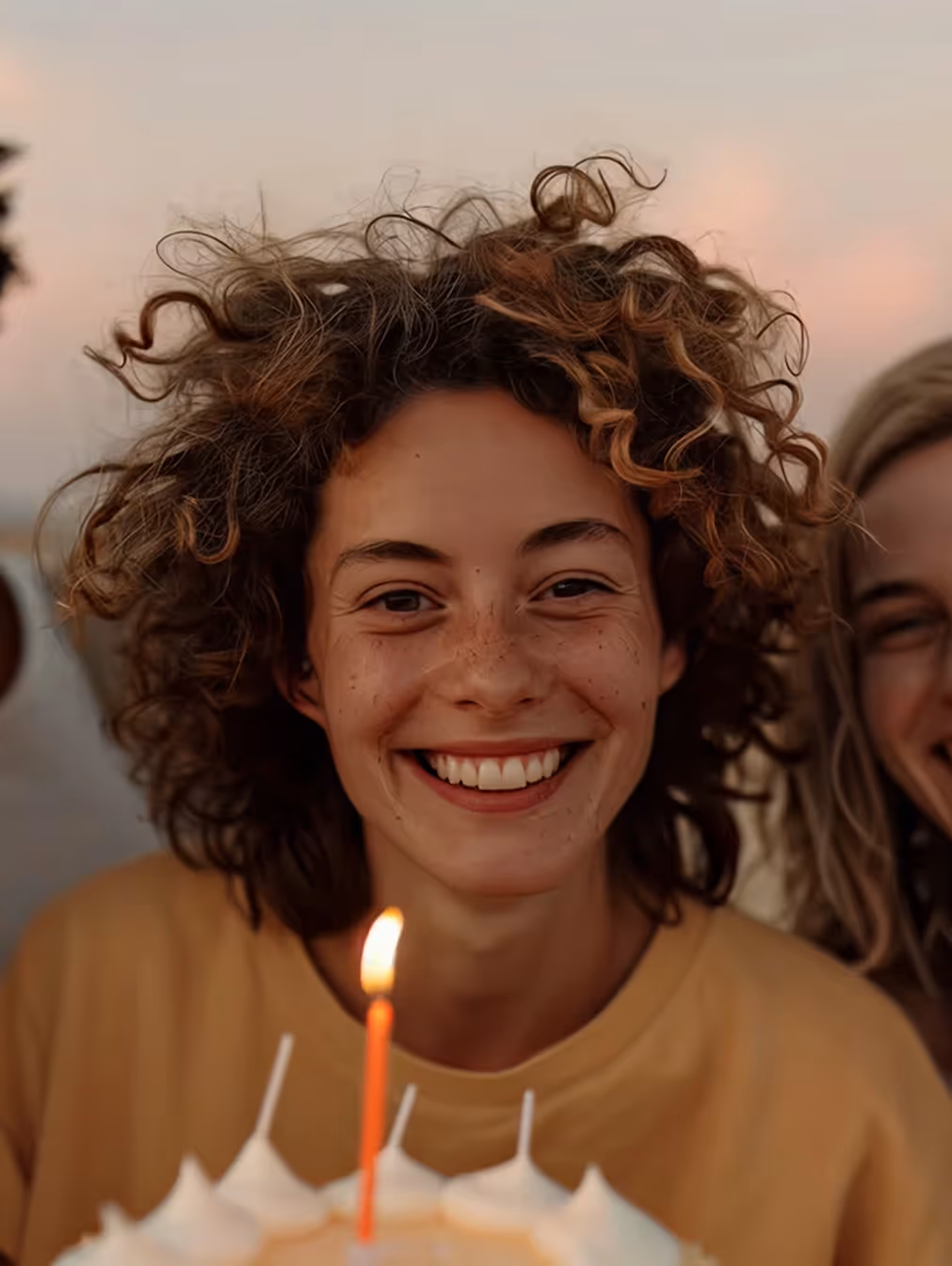 A person with curly hair smiling widely behind a birthday cake with a lit candle, joined by a friend in the background.