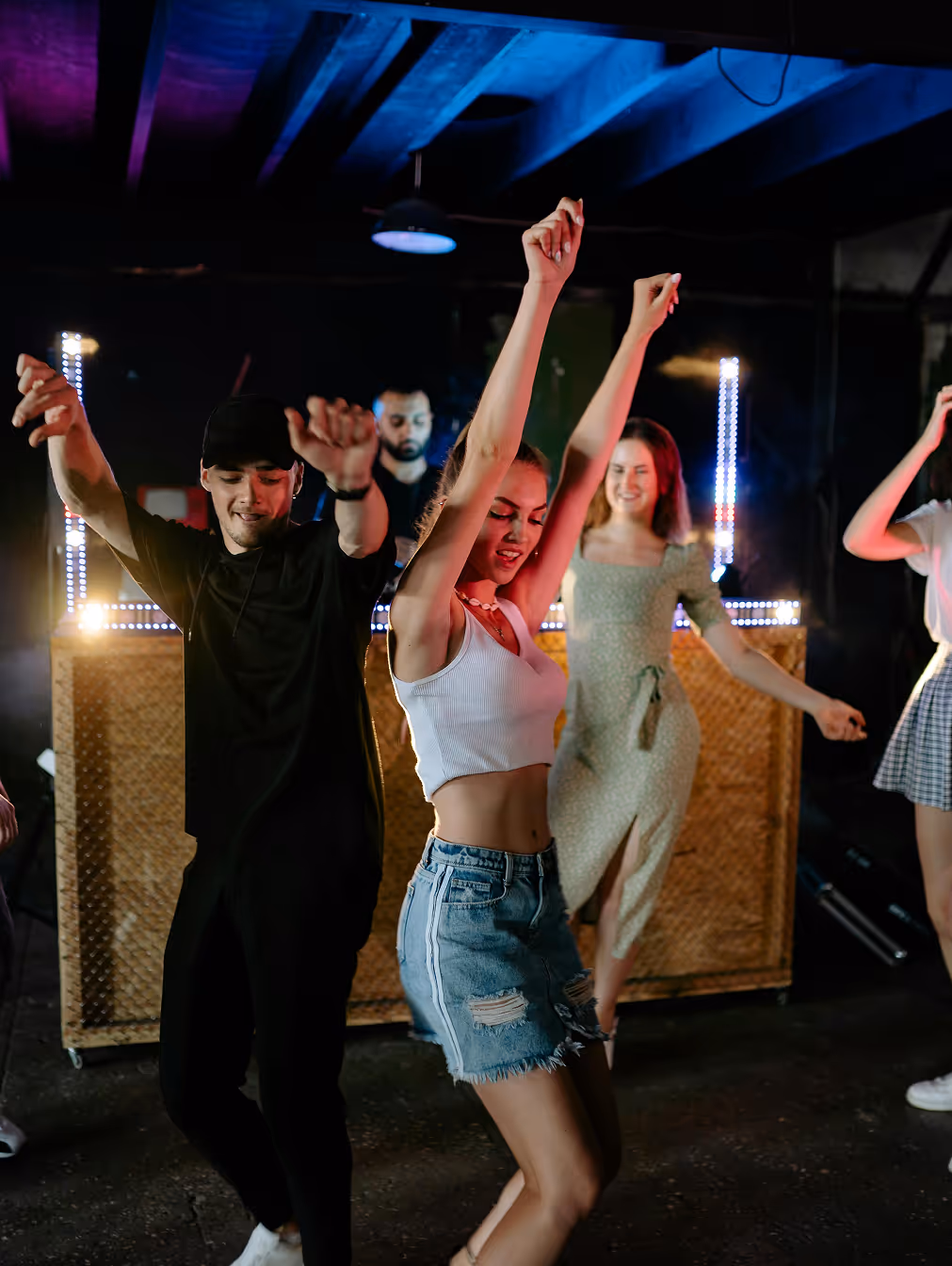 A group of people dancing energetically under bright lights in a club, with a DJ booth in the background.