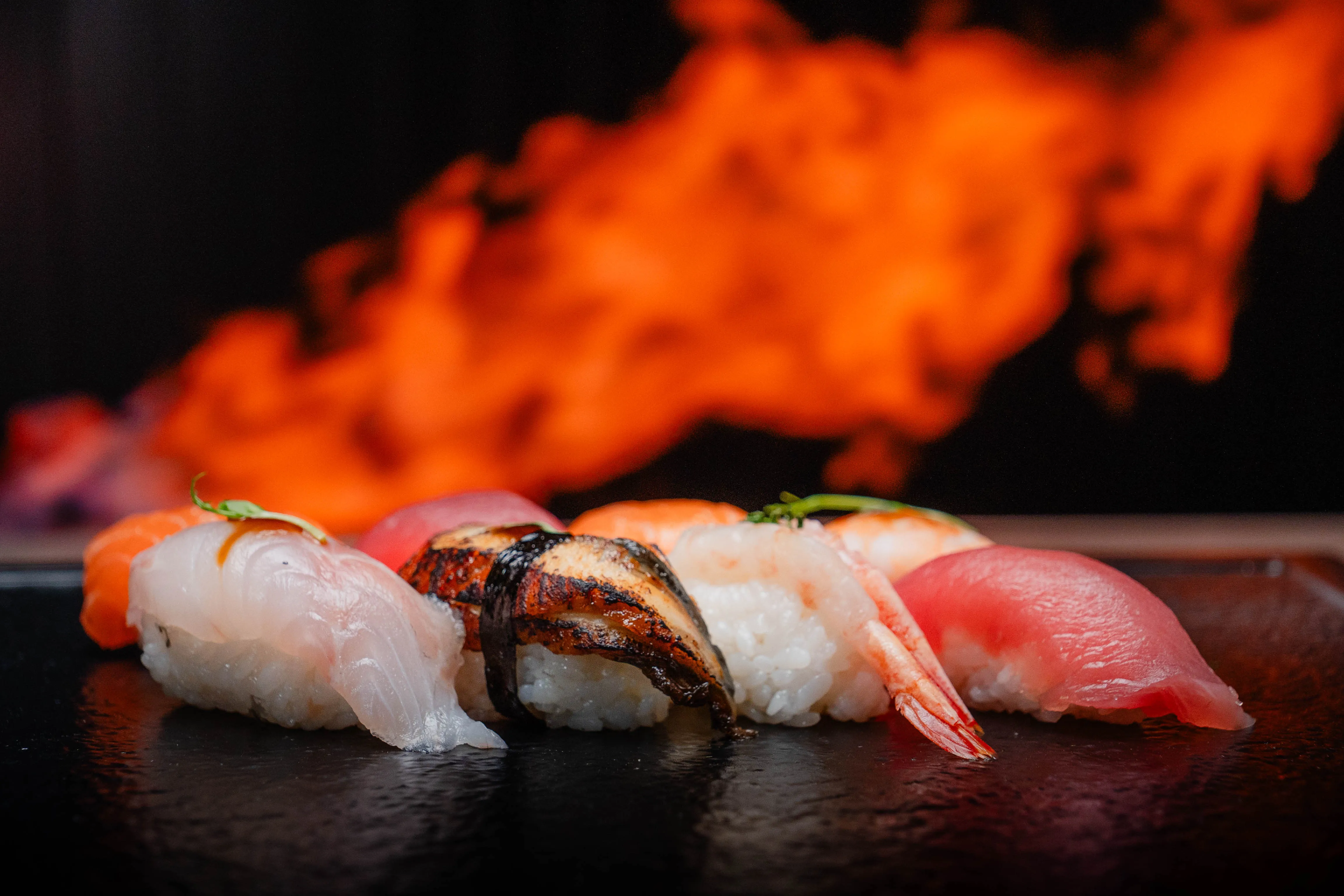 Nahaufnahme von verschiedenen Sushi-Stücken auf schwarzem Teller vor Flammen im Hintergrund.