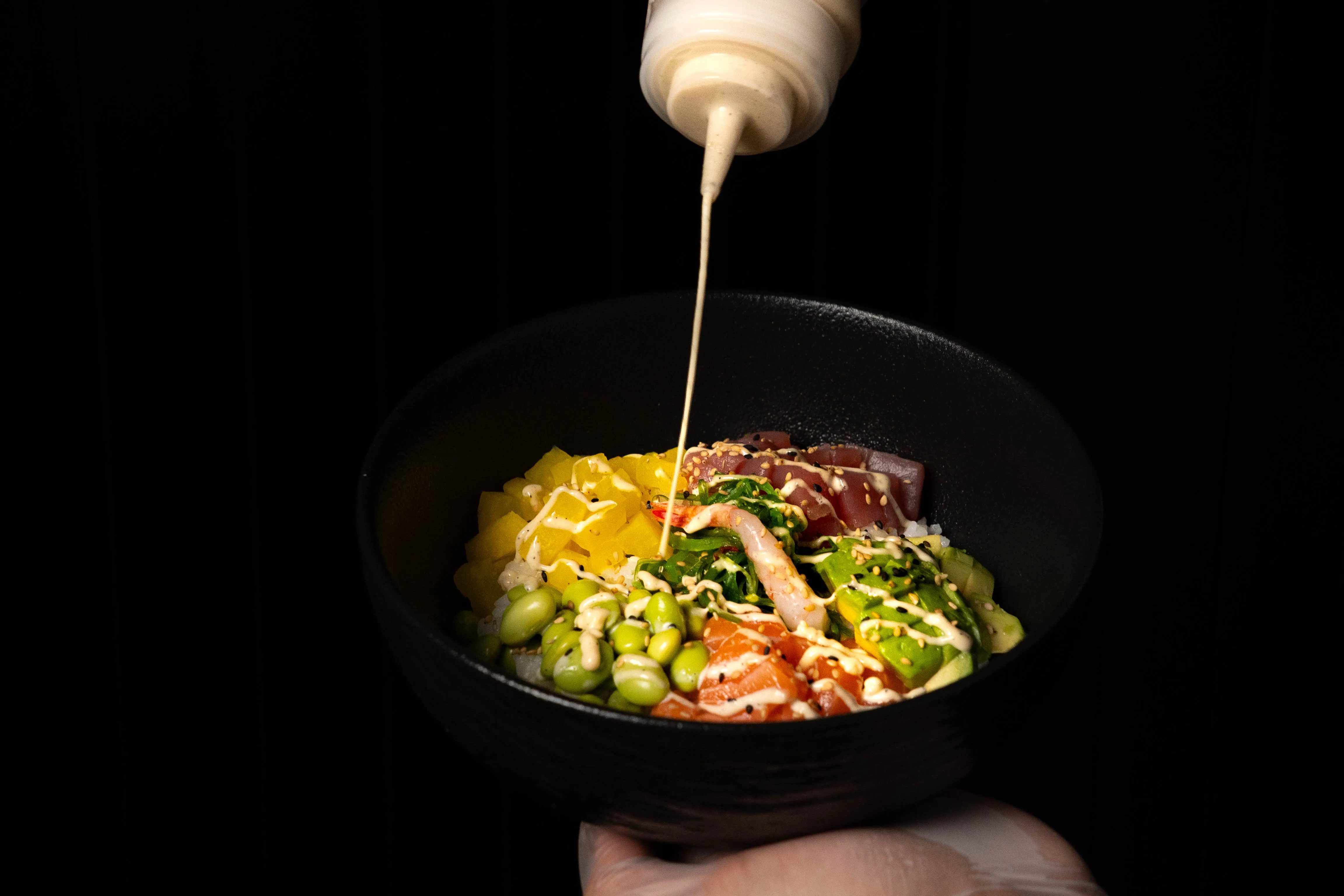 Schwarze Schüssel mit Sushi-Zutaten und Sesamsauce, die aus einer Flasche darüber geträufelt wird.