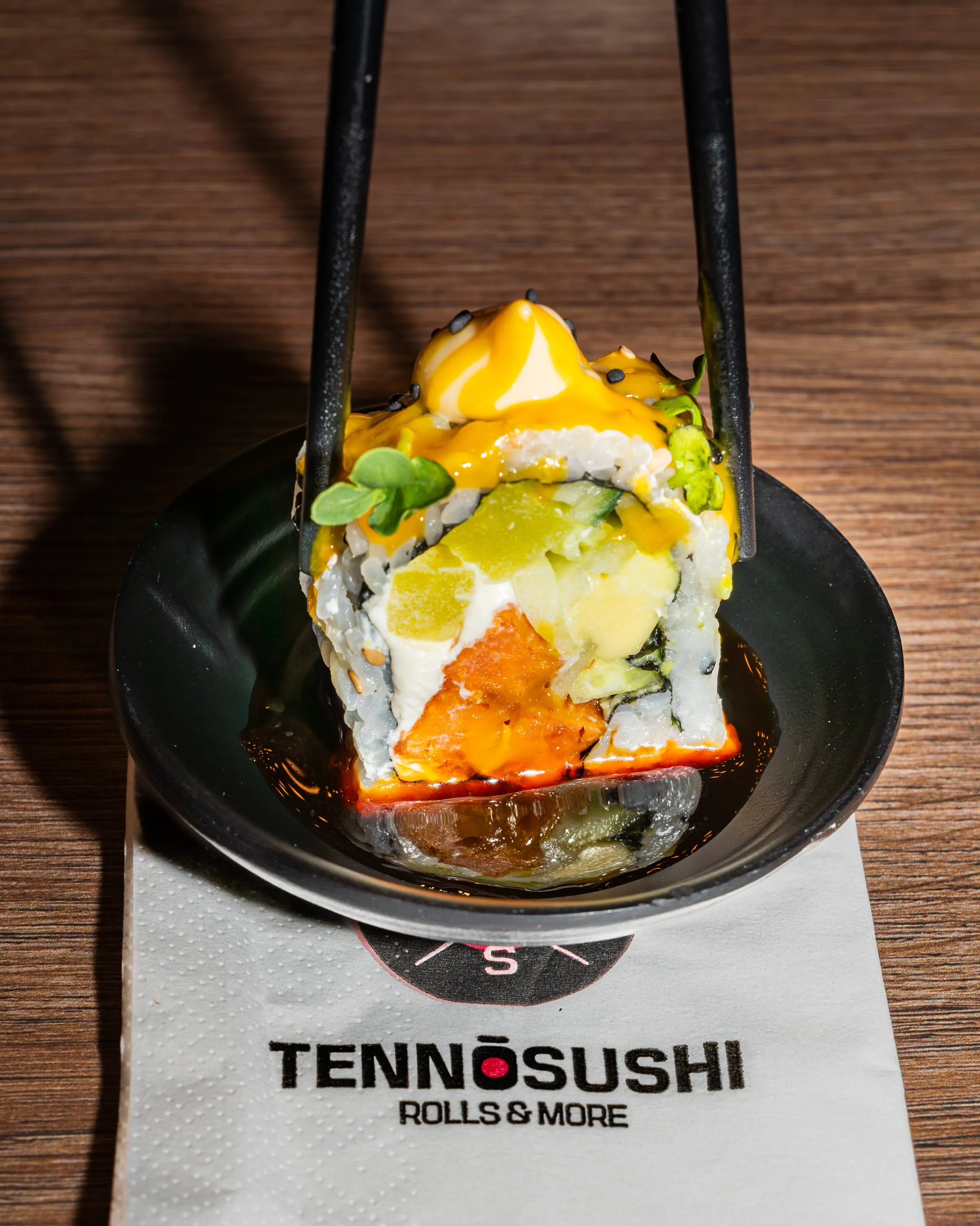 Sushirolle mit Mango, Avocado und Sauce in schwarzer Schale, gehalten von schwarzen Stäbchen.