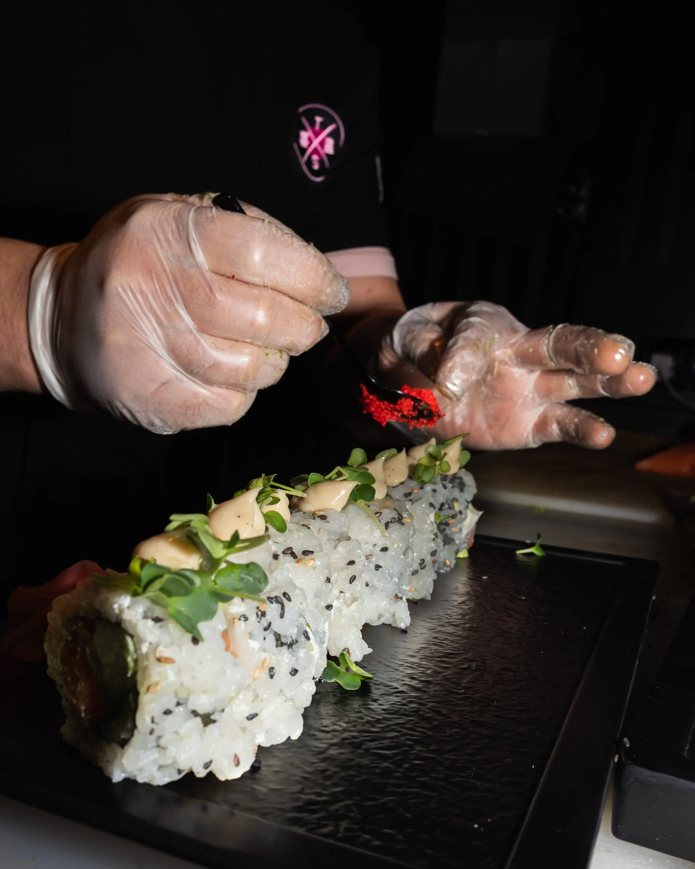 Eine Hand mit Handschuhen garniert Sushi mit kleinen roten Fischrogen auf einem schwarzen Tablett.