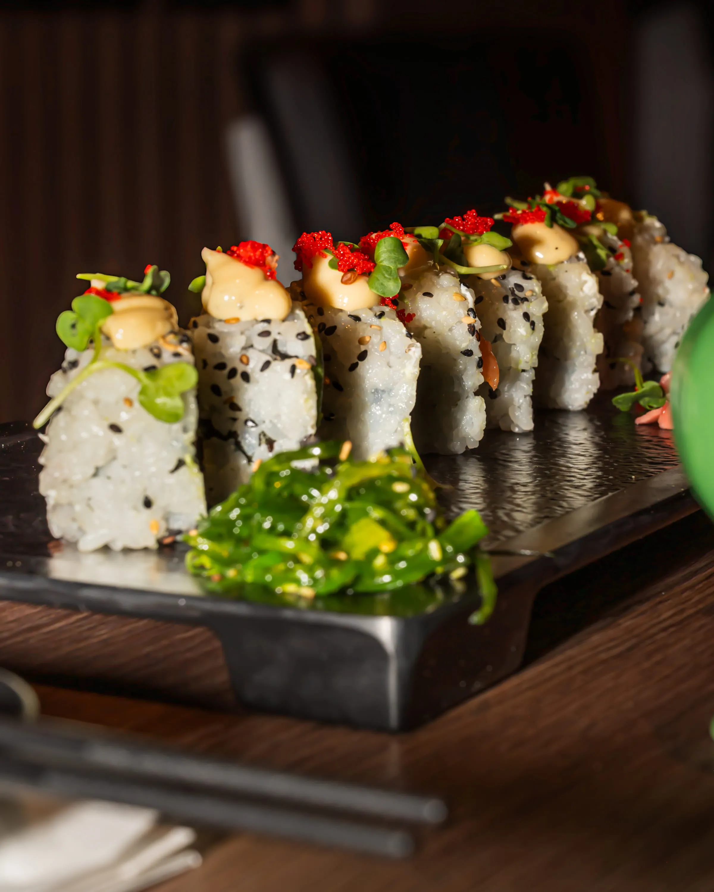 Reihe von Sushi-Rollen mit Sesam, Mayonnaise und roten Fischrogen, serviert auf schwarzem Brett mit Algensalat.