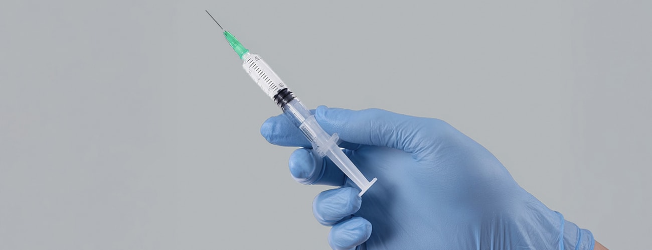 A hand wearing a blue latex glove holding a filled syringe with a green needle.