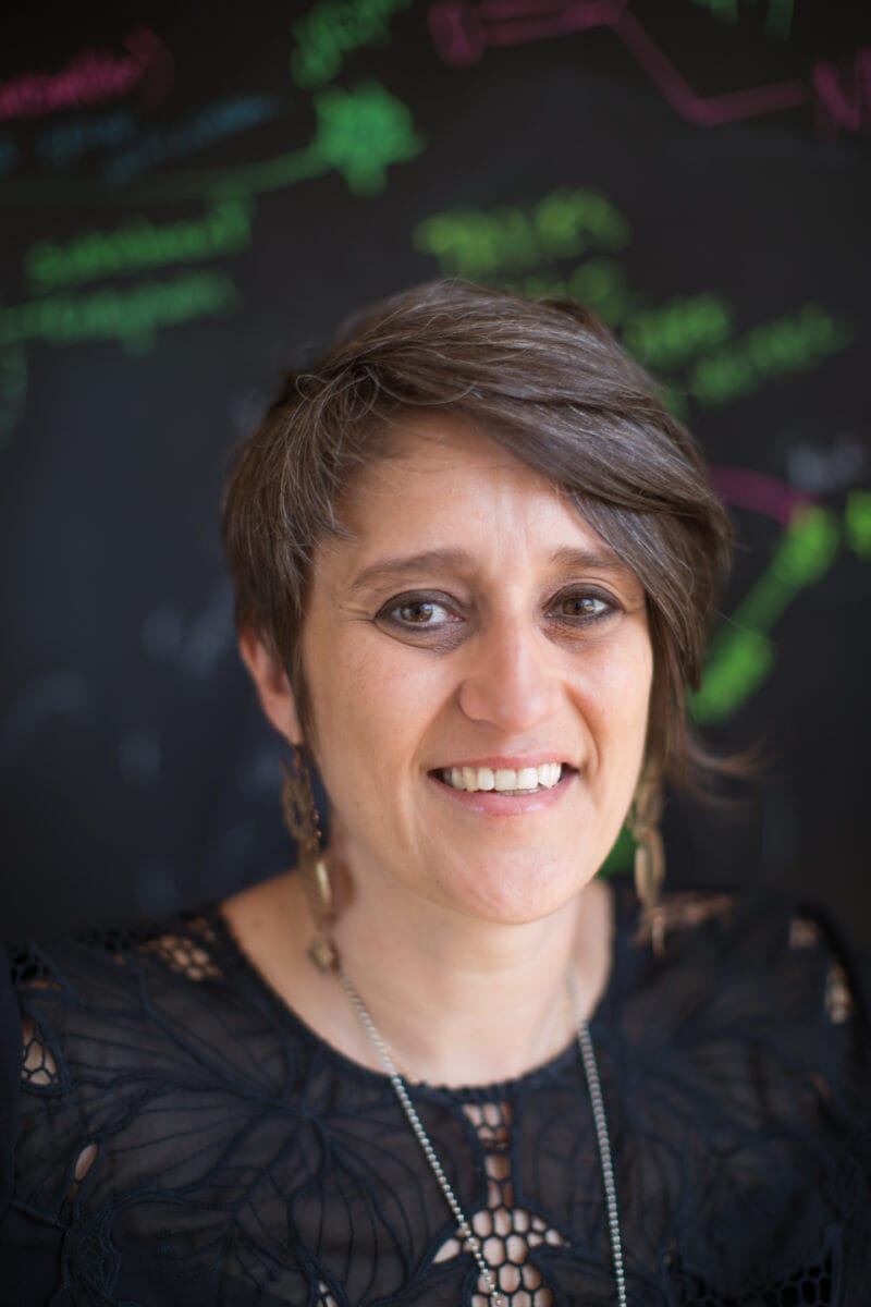 Gul Dolen standing in front of a blackboard with colorful writing.