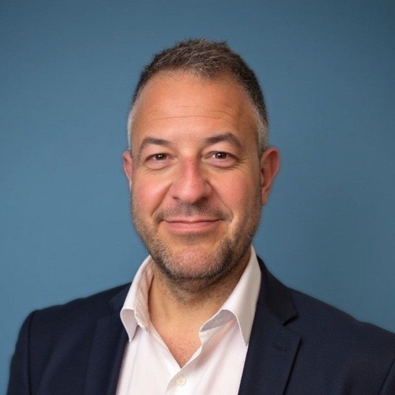 Simeon Schnapper wearing a white shirt and dark blazer against a blue background.