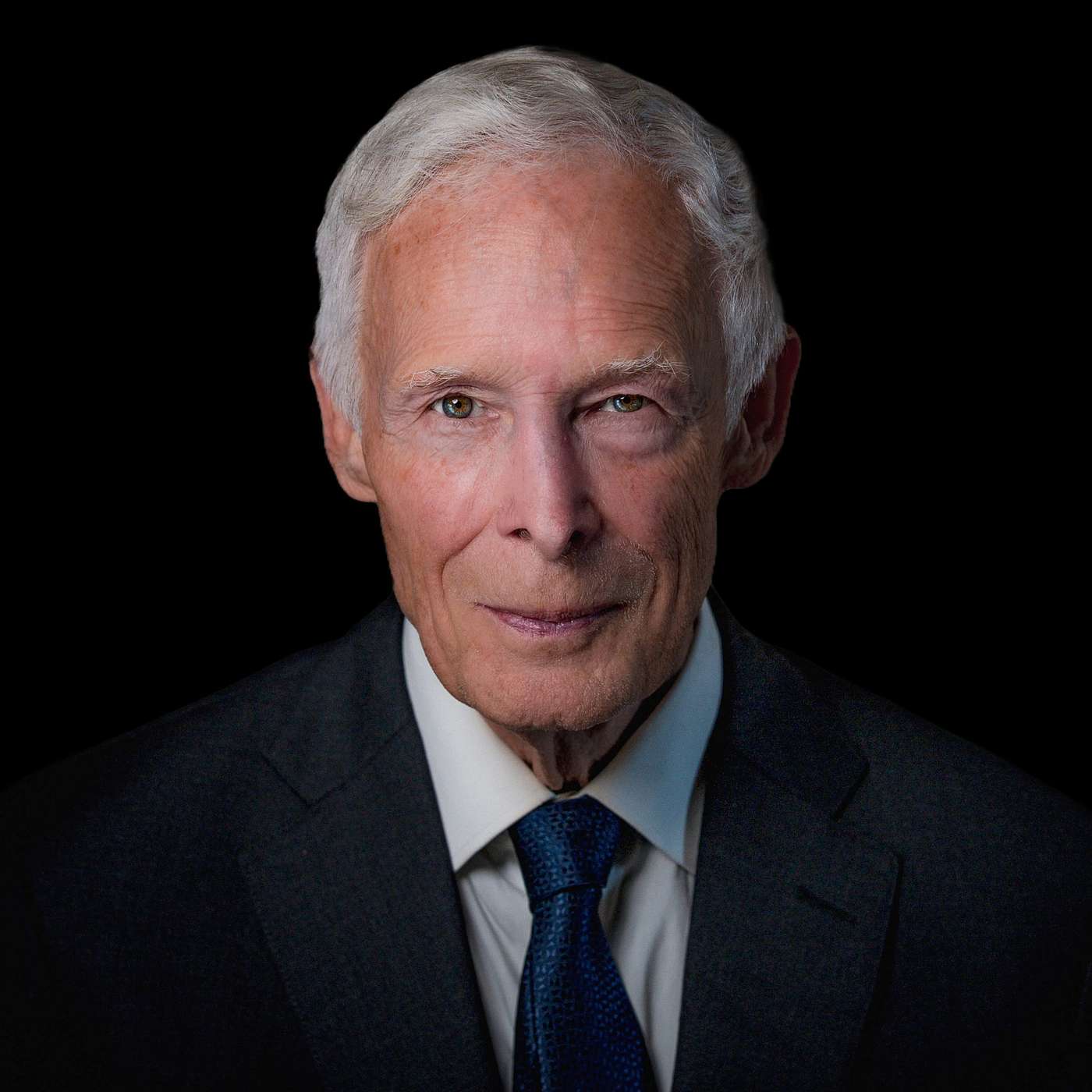 Portrait of Leonard Pickard wearing a dark suit, white shirt, and blue tie against a black background.