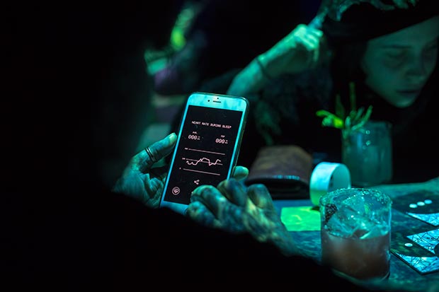 Person holding a smartphone displaying a heart rate monitoring app in a dimly lit setting with another person blurred in the background.