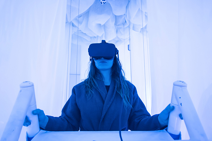 Person wearing a virtual reality headset and holding white handles inside a blue-lit immersive environment.