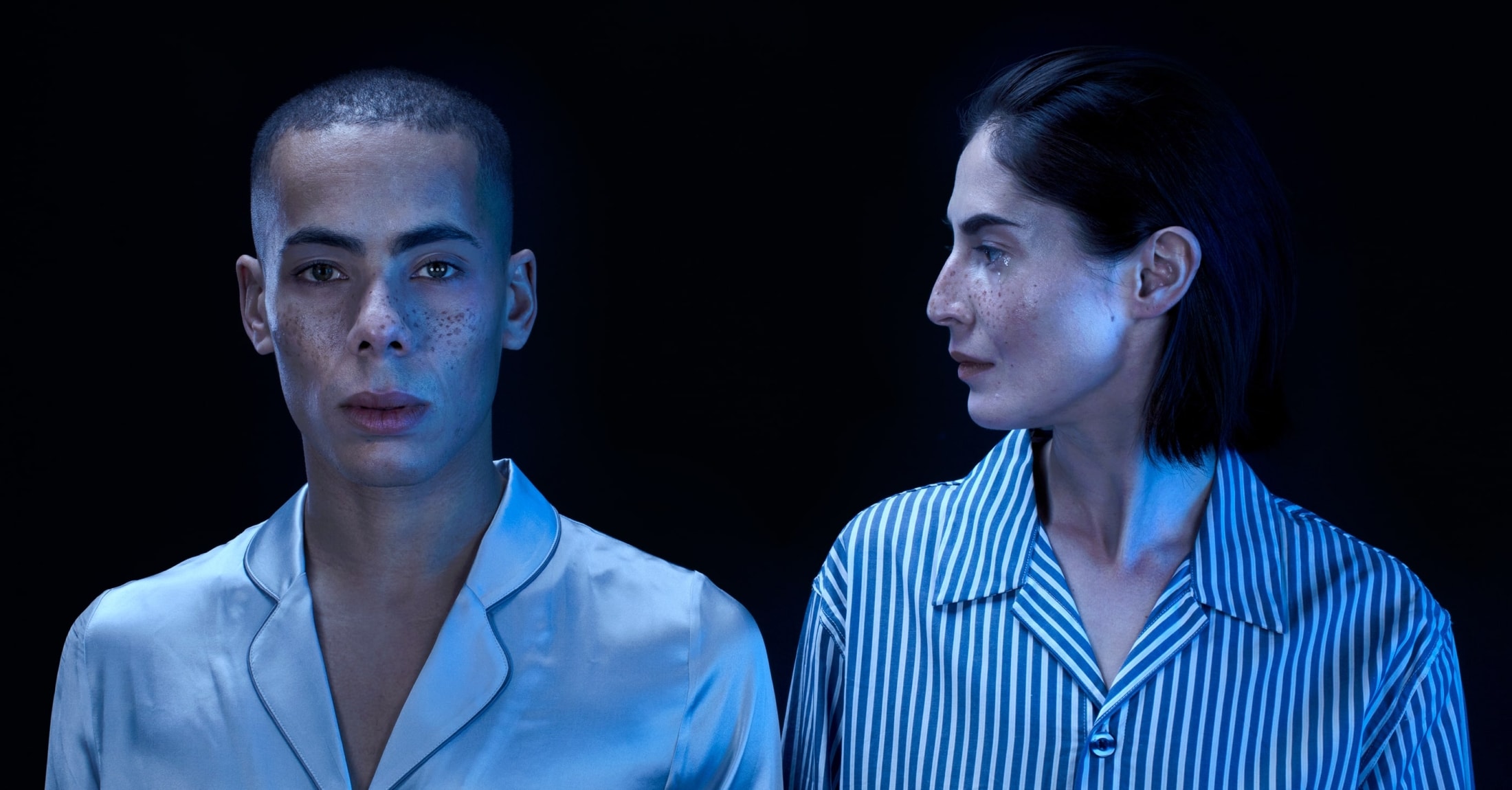 Portrait of a man in a light blue satin shirt looking forward and a woman in a blue striped shirt looking to the right, both with freckles and illuminated by blue light against a dark background.