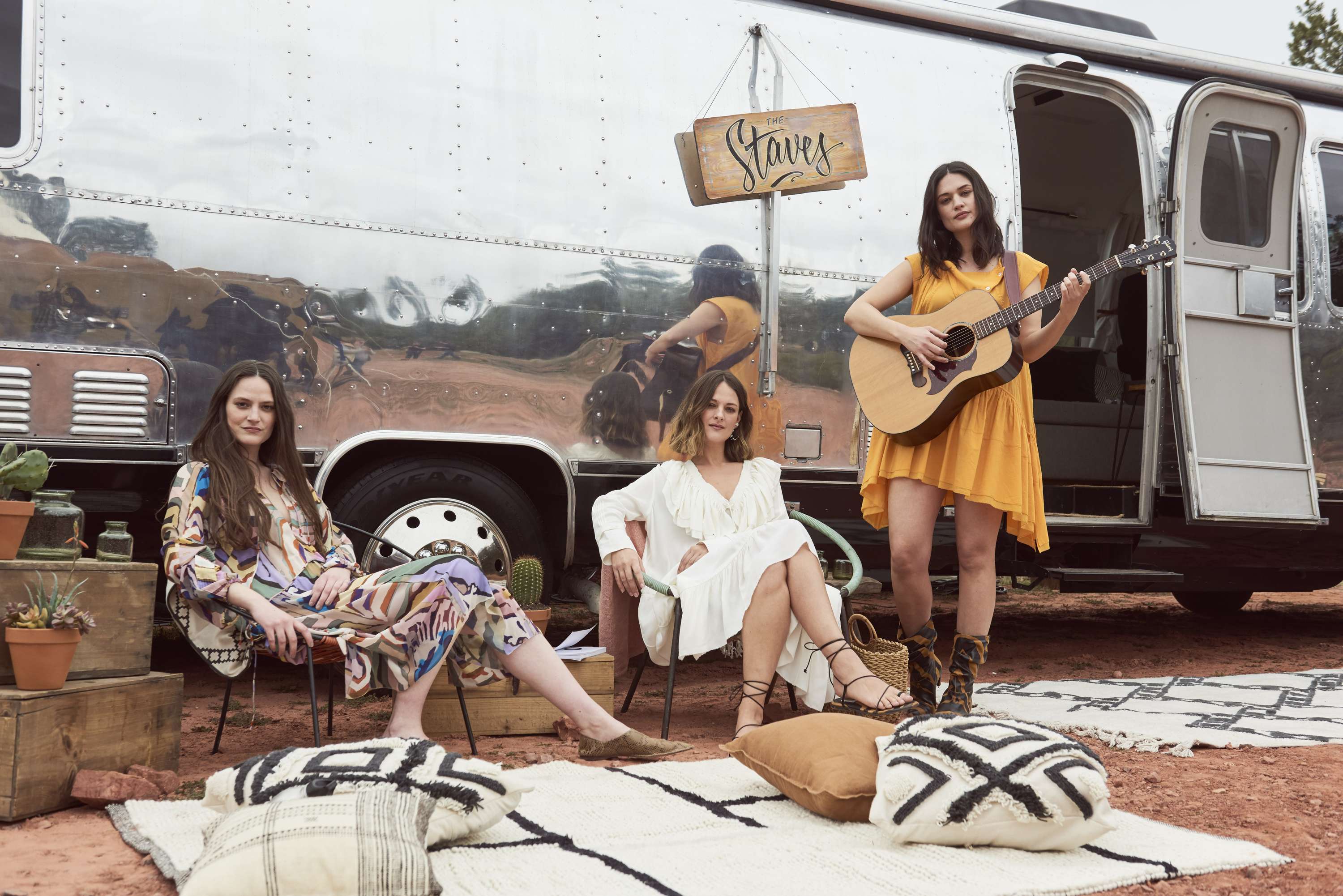 Three women near a silver trailer; one plays guitar in a yellow dress, two others sit on chairs surrounded by cushions and plants.