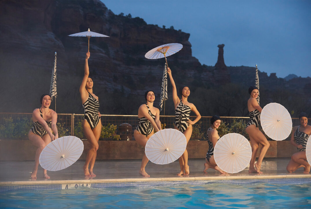 Seven women in zebra-striped swimsuits performing a synchronized routine by a pool holding white parasols at dusk.