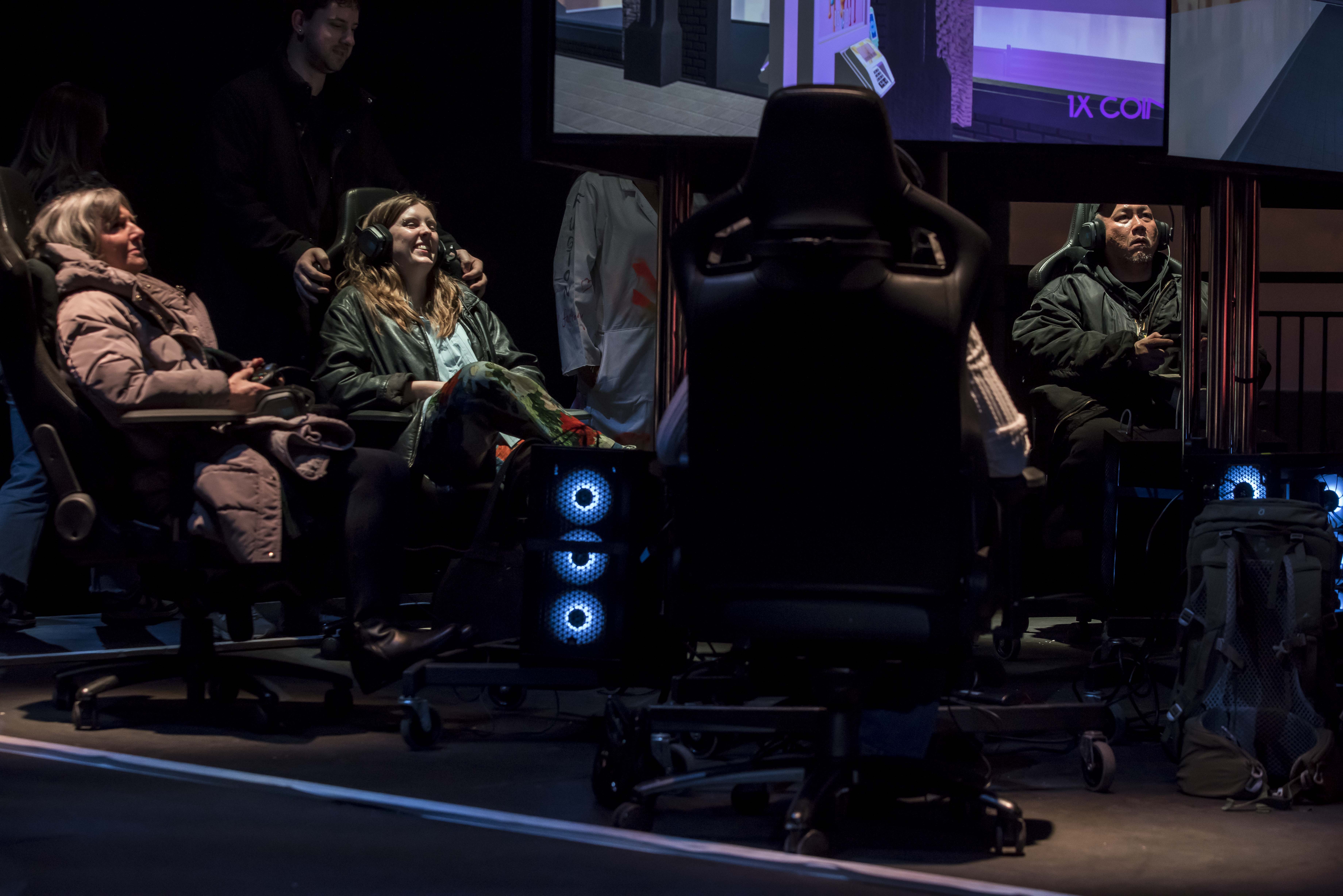 People seated in gaming chairs wearing headphones, focused on screens in a dark room with ambient blue lighting.