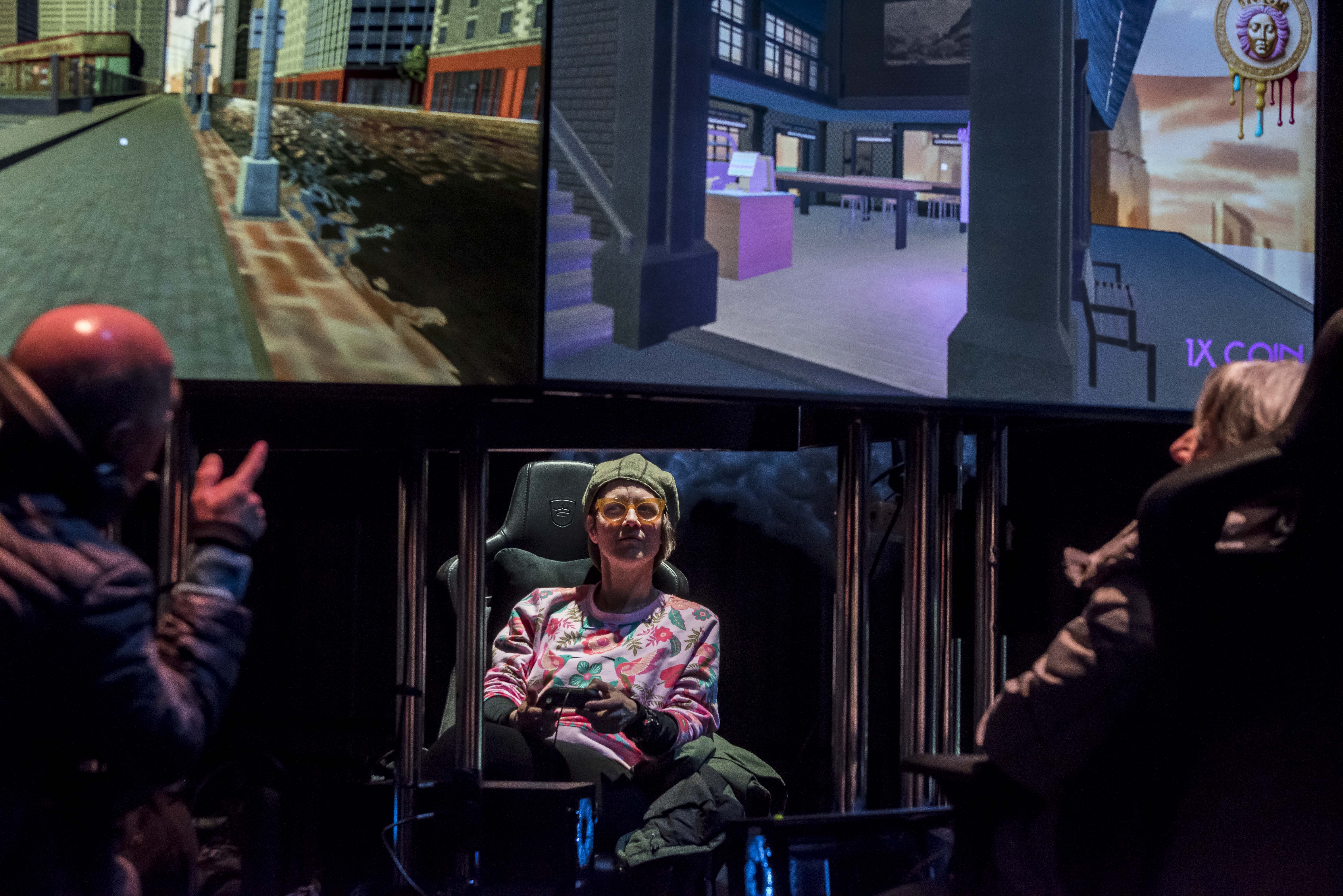 Person wearing glasses and a hat plays a video game seated in a gaming chair with two large screens displaying virtual environments behind them.