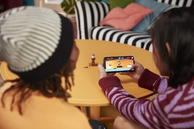 Two people sitting at a yellow table; one holds a smartphone showing a game with small character figurines on the table.