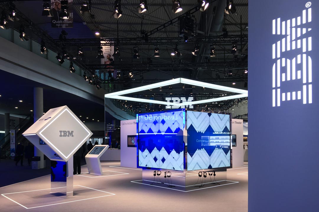 IBM exhibition booth with illuminated logos, digital screens, and interactive displays under a ceiling with numerous lights.