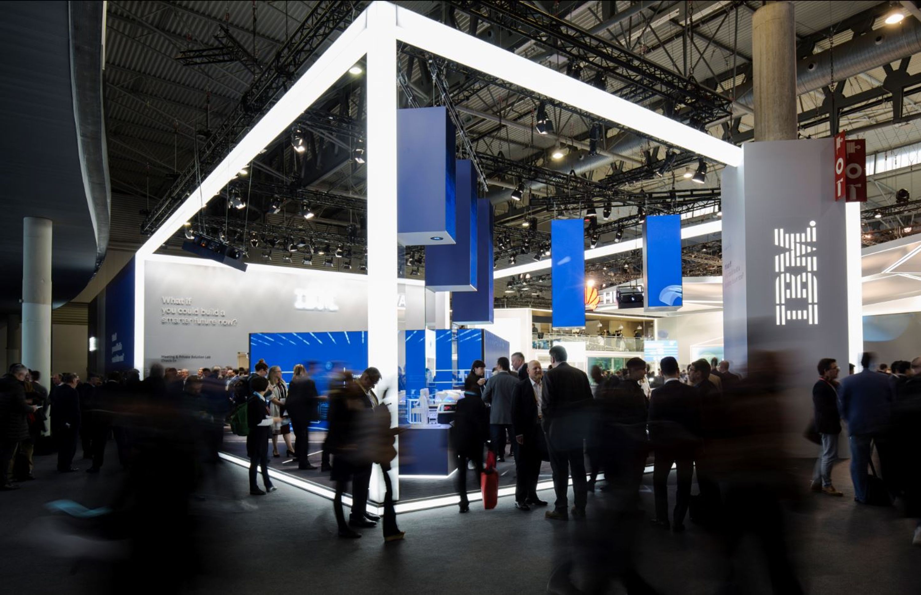 Busy IBM exhibition booth with illuminated white frame and blue hanging panels at a technology event.