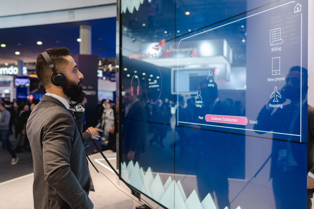 Man in a suit wearing headphones interacts with a large touch screen displaying customer satisfaction data at a tech event.