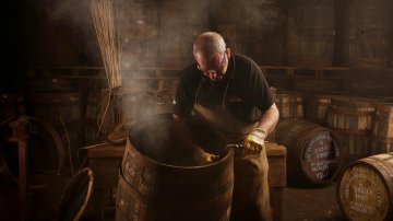 Glenfiddich - man stirring steaming contents inside a wooden barrel in a dimly lit distillery with barrels in the background.