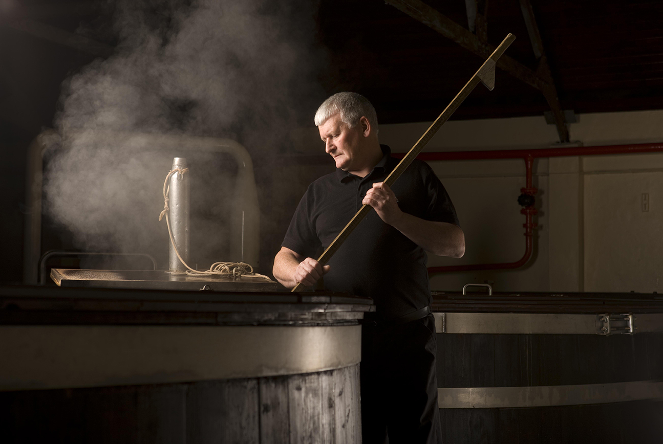 Glenfiddich - a man with gray hair stirring a steaming fermentation vat with a long wooden paddle inside a dimly lit distillery.