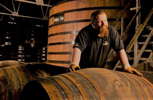 Glendfiddich craftsman - man with a beard leaning on large wooden barrels in a dimly lit warehouse.