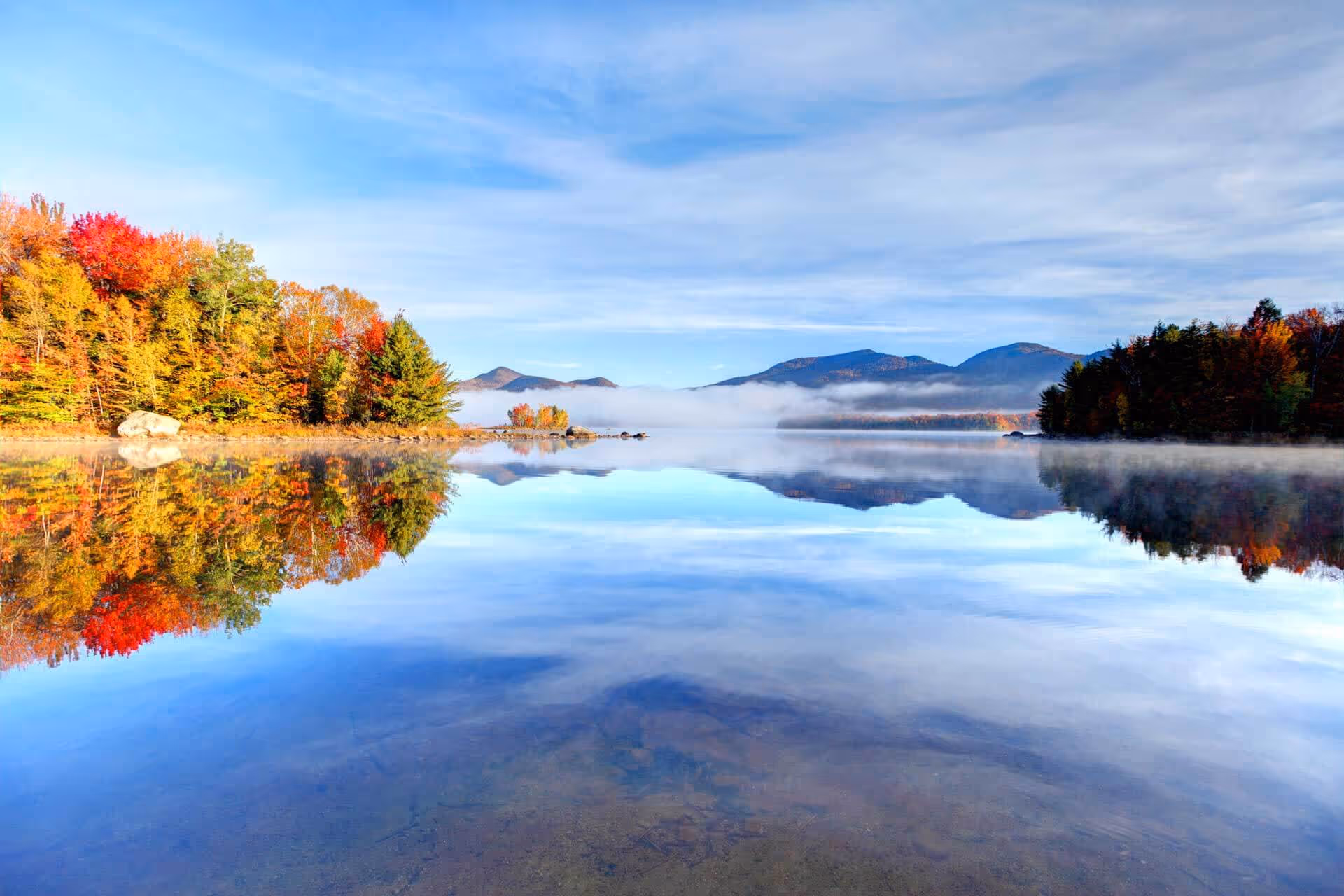 Calm lake reflecting colorful autumn trees and distant mountains under a partly cloudy sky.