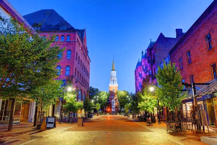 Evening view of a pedestrian street lined with trees and brick buildings, centered on a white church with a tall golden steeple under a clear blue sky.