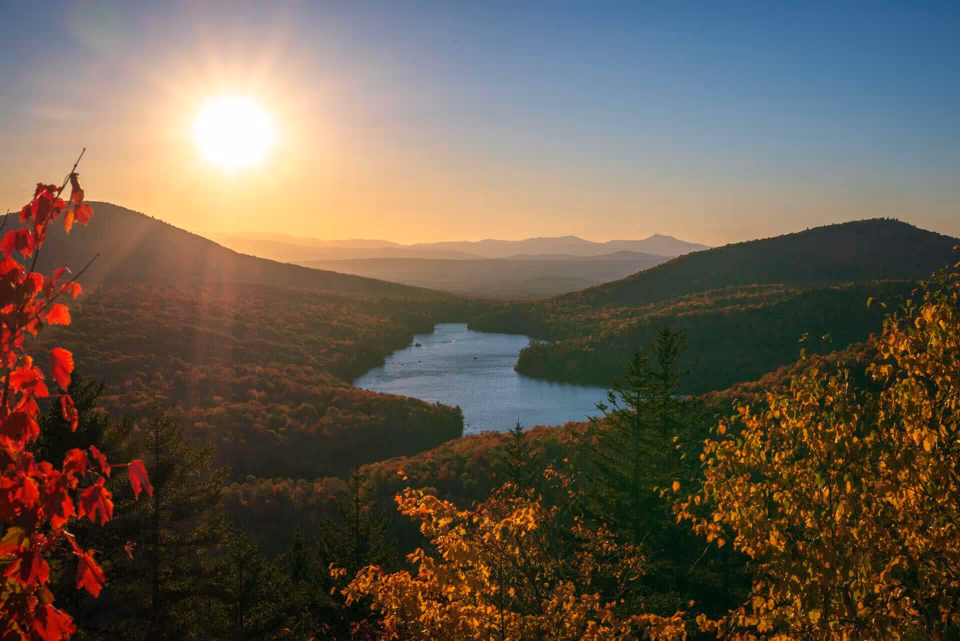 Sunset over a lake surrounded by forested mountains with colorful autumn foliage.