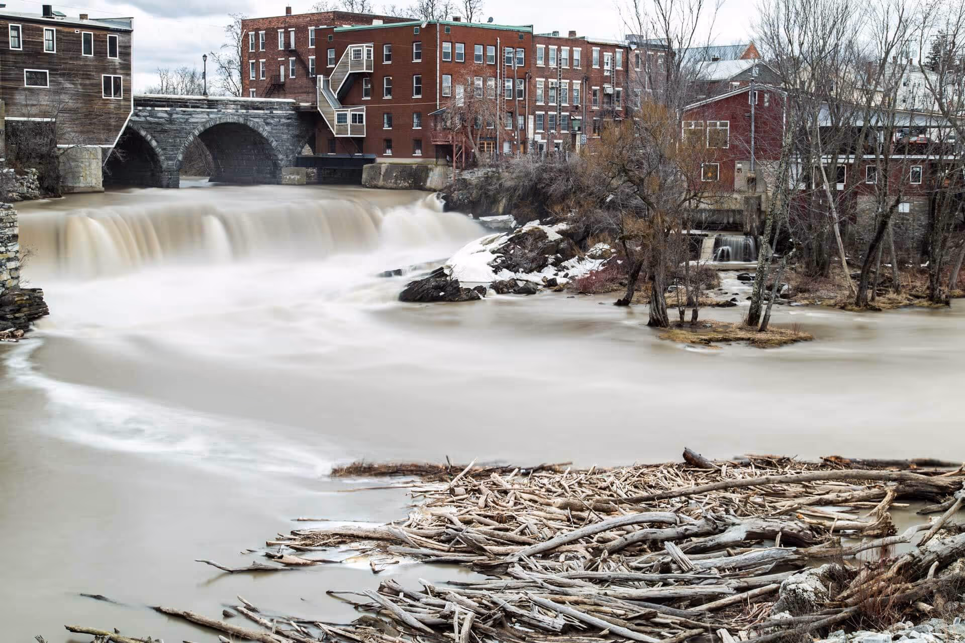 Calm river with driftwood in the foreground, a stone arch bridge, waterfall, and red brick buildings in a small town setting.